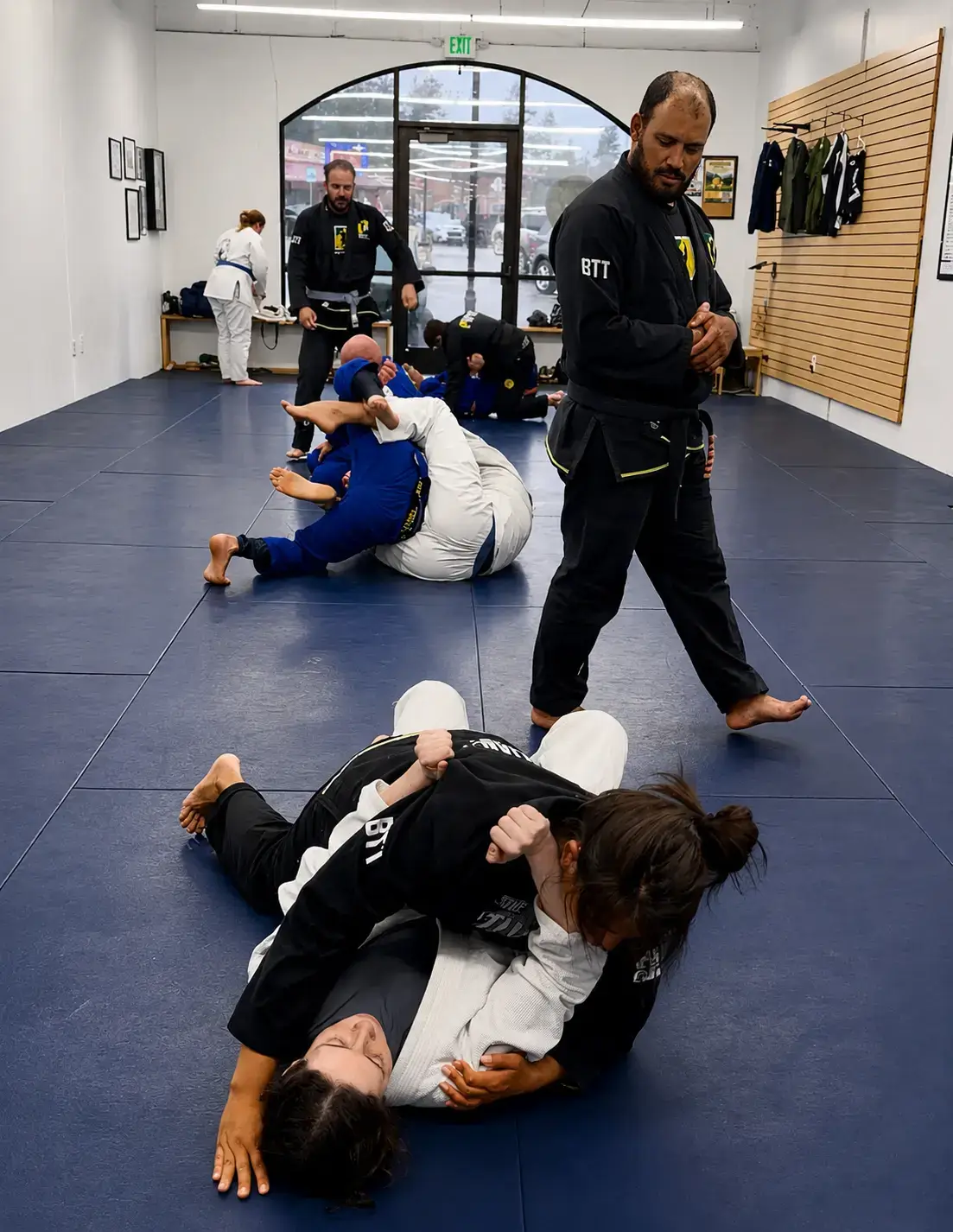 Brazilian Jiu-Jitsu students in gis practice grappling on blue mats while an instructor observes them.