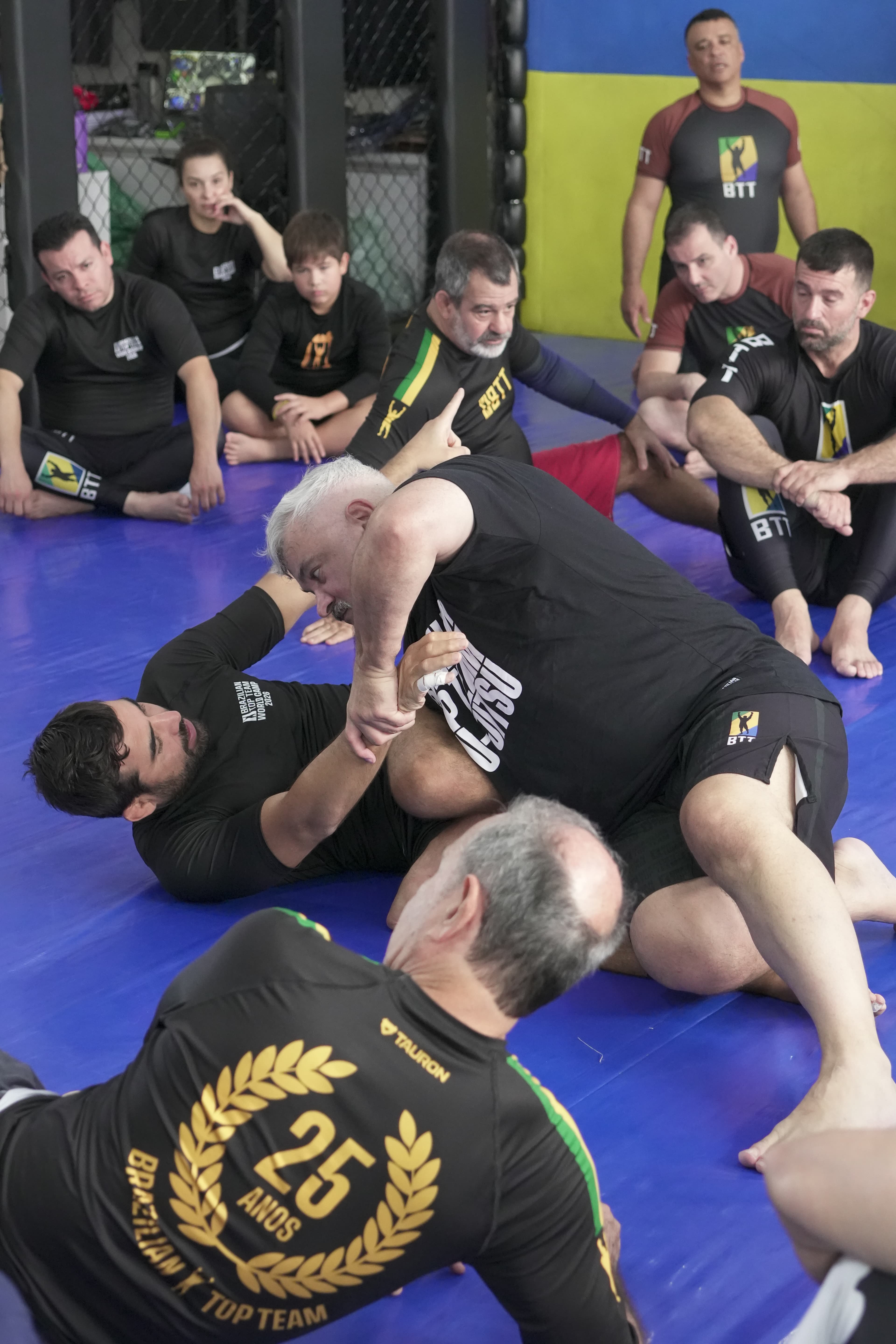 Two men demonstrate a Brazilian Jiu-Jitsu grappling technique on a blue mat while others watch.