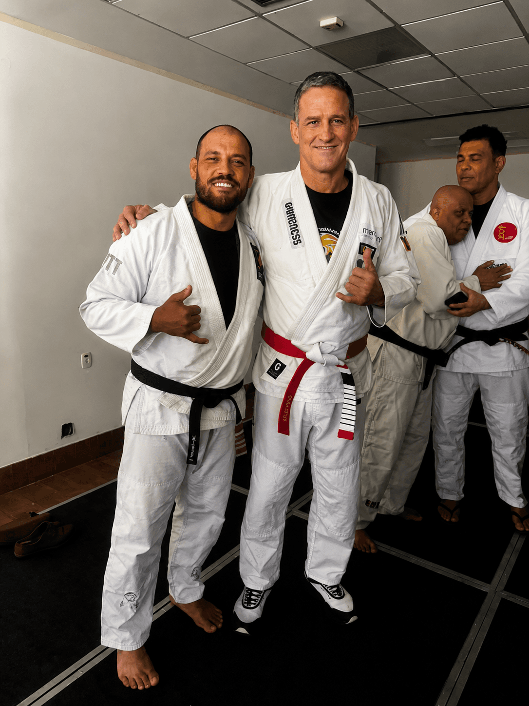 Two smiling men in white martial arts gis pose together, making shaka hand signs.