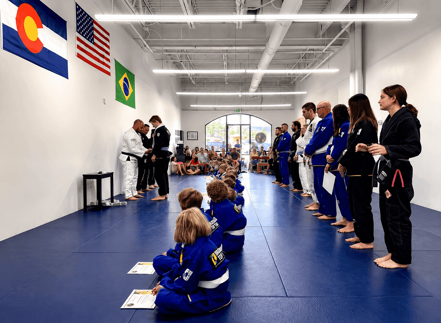 Students watch a martial arts belt promotion ceremony on blue mats in a bright gym.