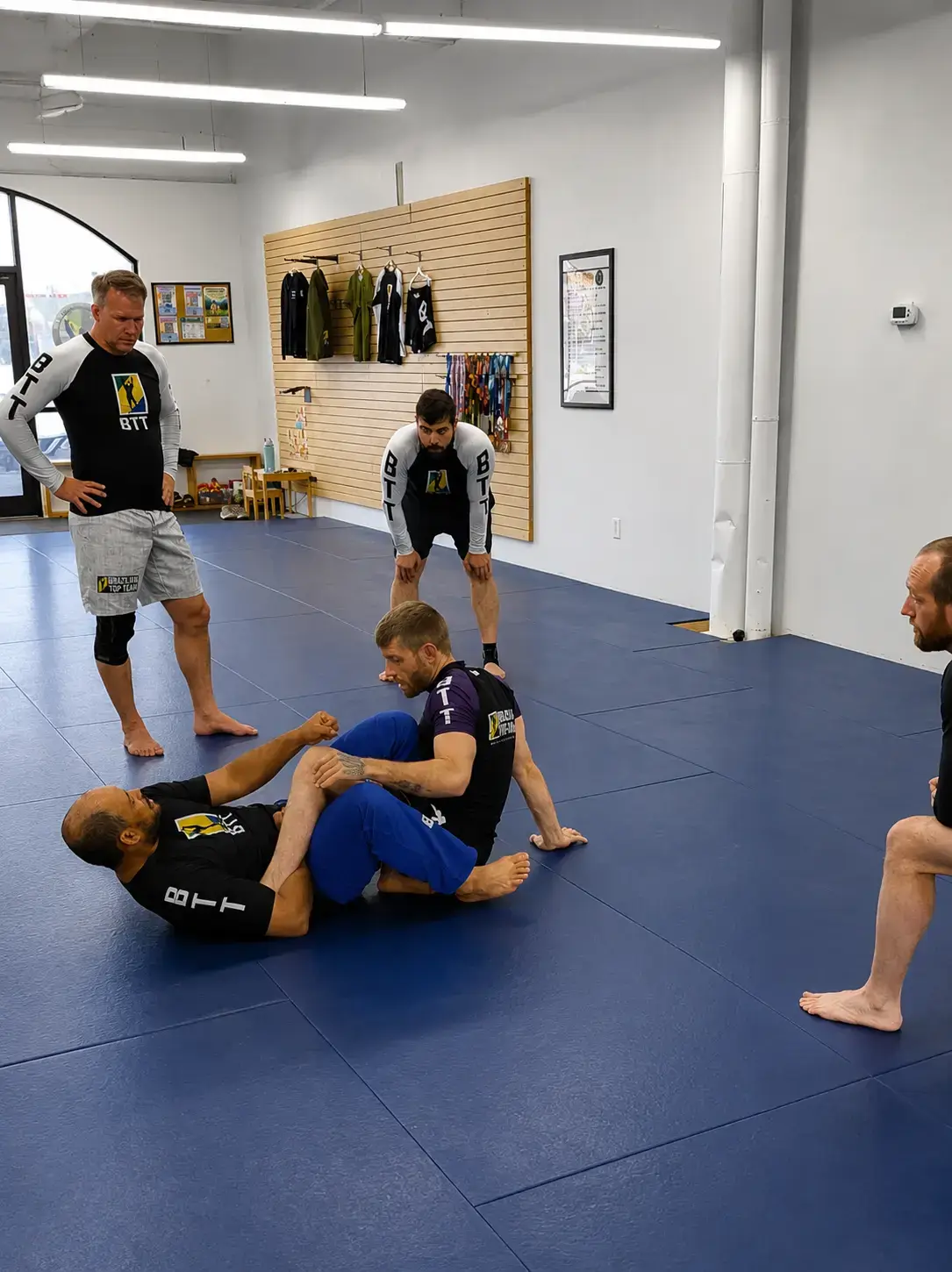 Brazilian Jiu-Jitsu practitioners demonstrate a leg lock technique on blue mats while others observe.
