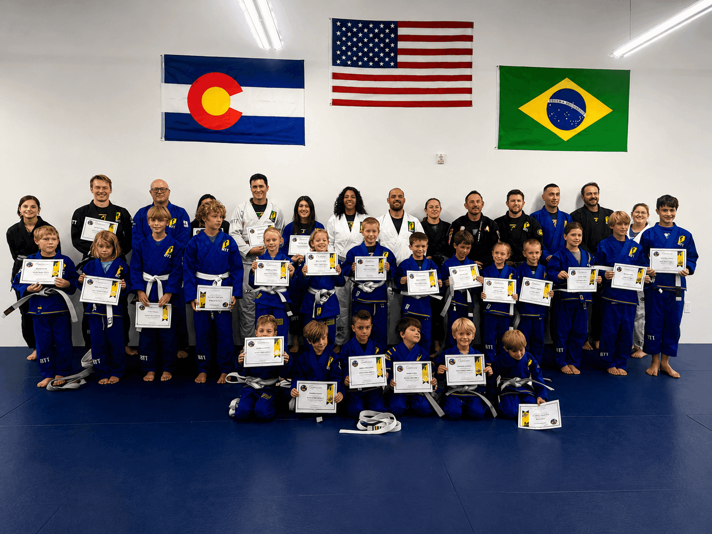 Martial arts students in blue gis hold certificates under Colorado, US, and Brazilian flags.