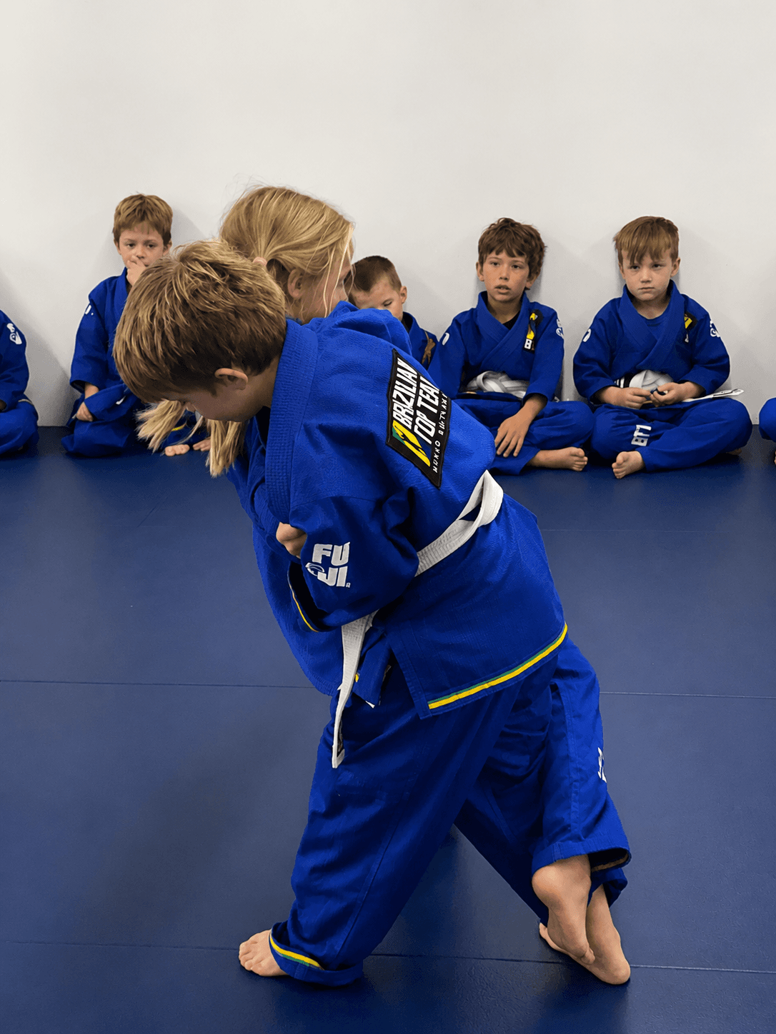Two children in blue gis practice grappling on a mat while other students watch.