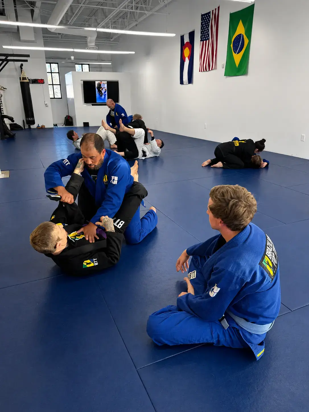 Brazilian Jiu-Jitsu students in blue and black gis practice grappling techniques on blue gym mats.
