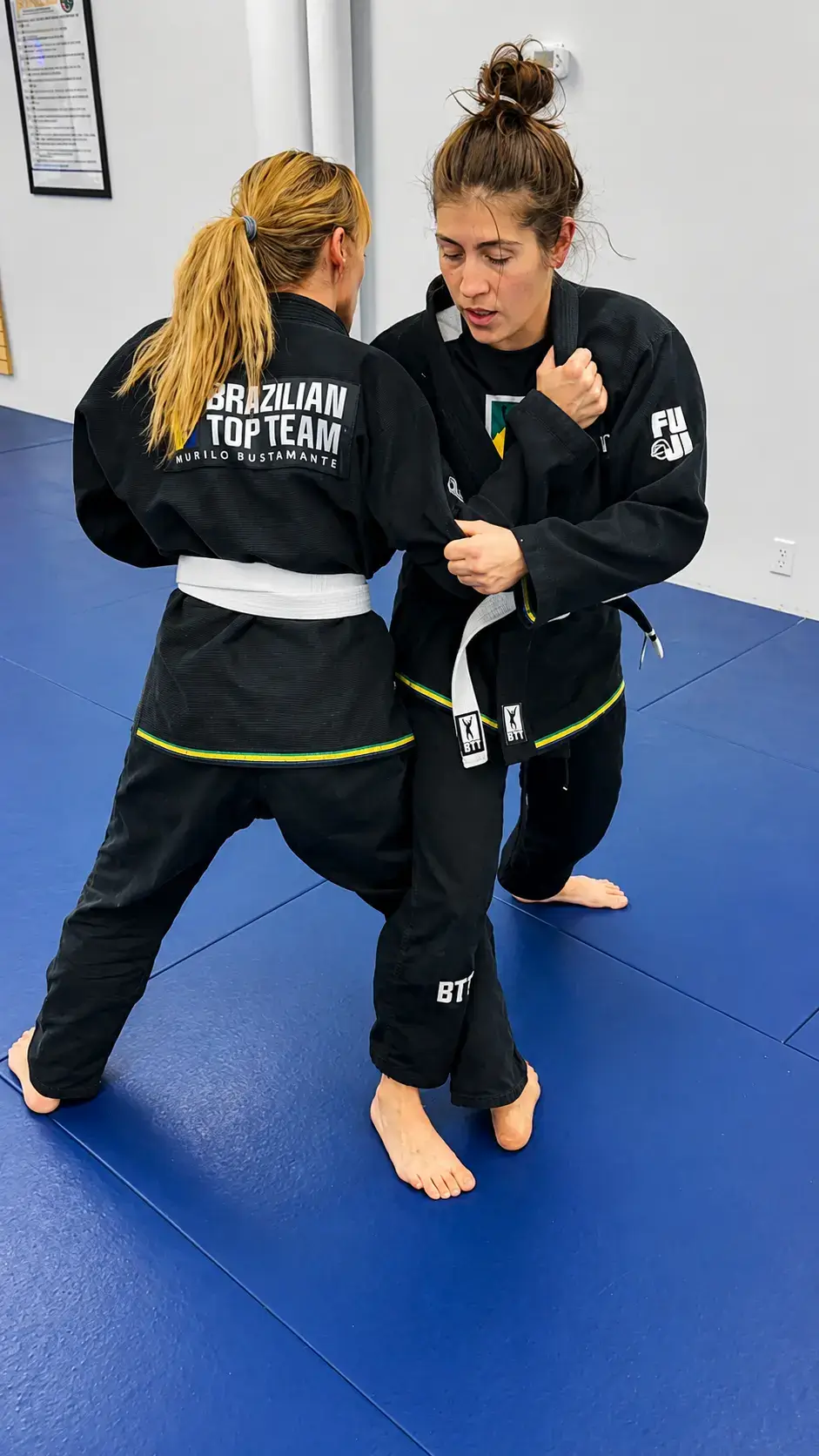 Two women in black gis practice Brazilian Jiu-Jitsu grappling techniques on a blue mat.