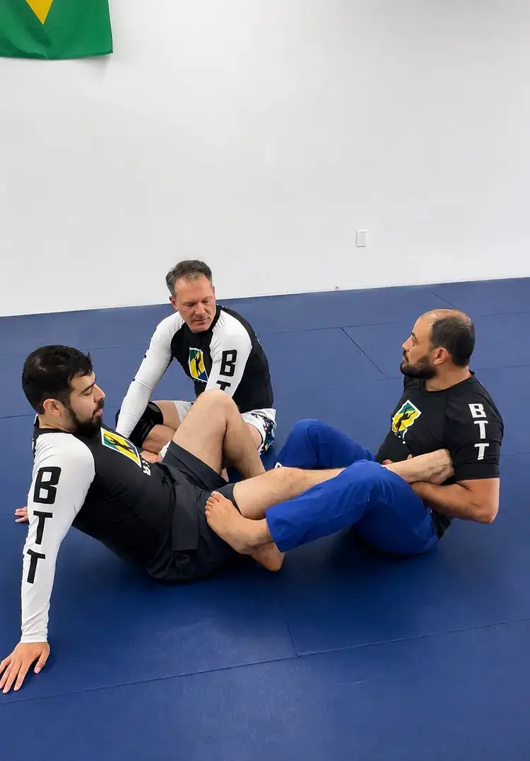 BJJ practitioners in branded rash guards demonstrate a leg lock technique on a blue mat.