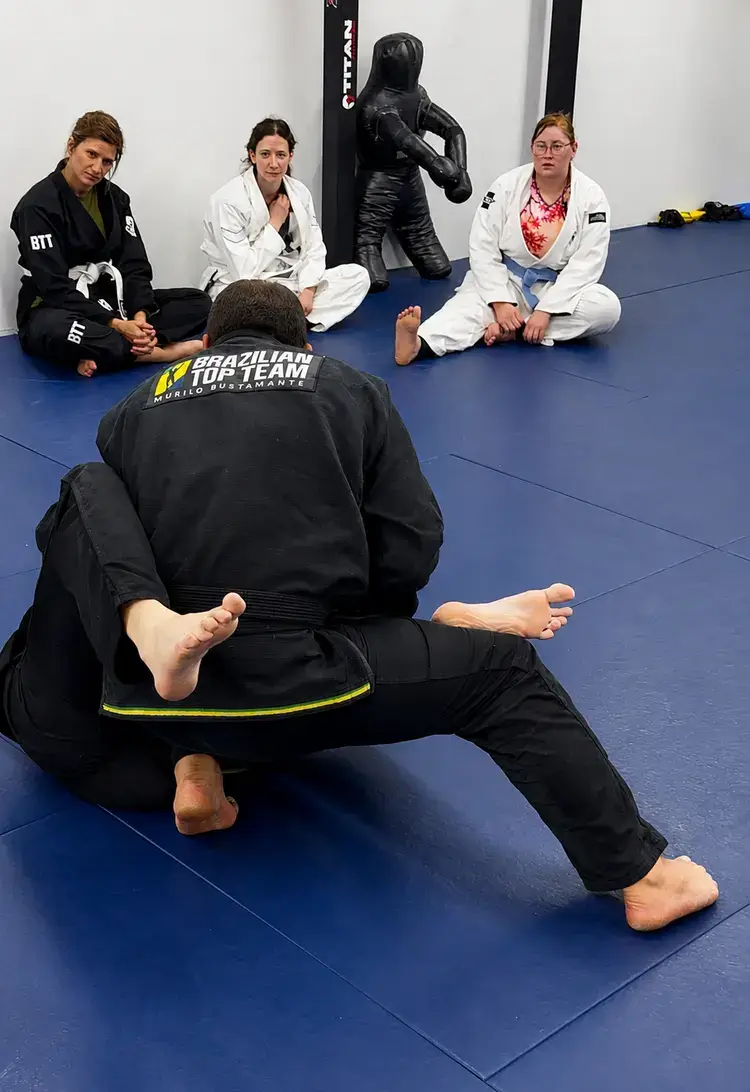 Instructor in black gi demonstrates BJJ technique to three students watching on blue mats.