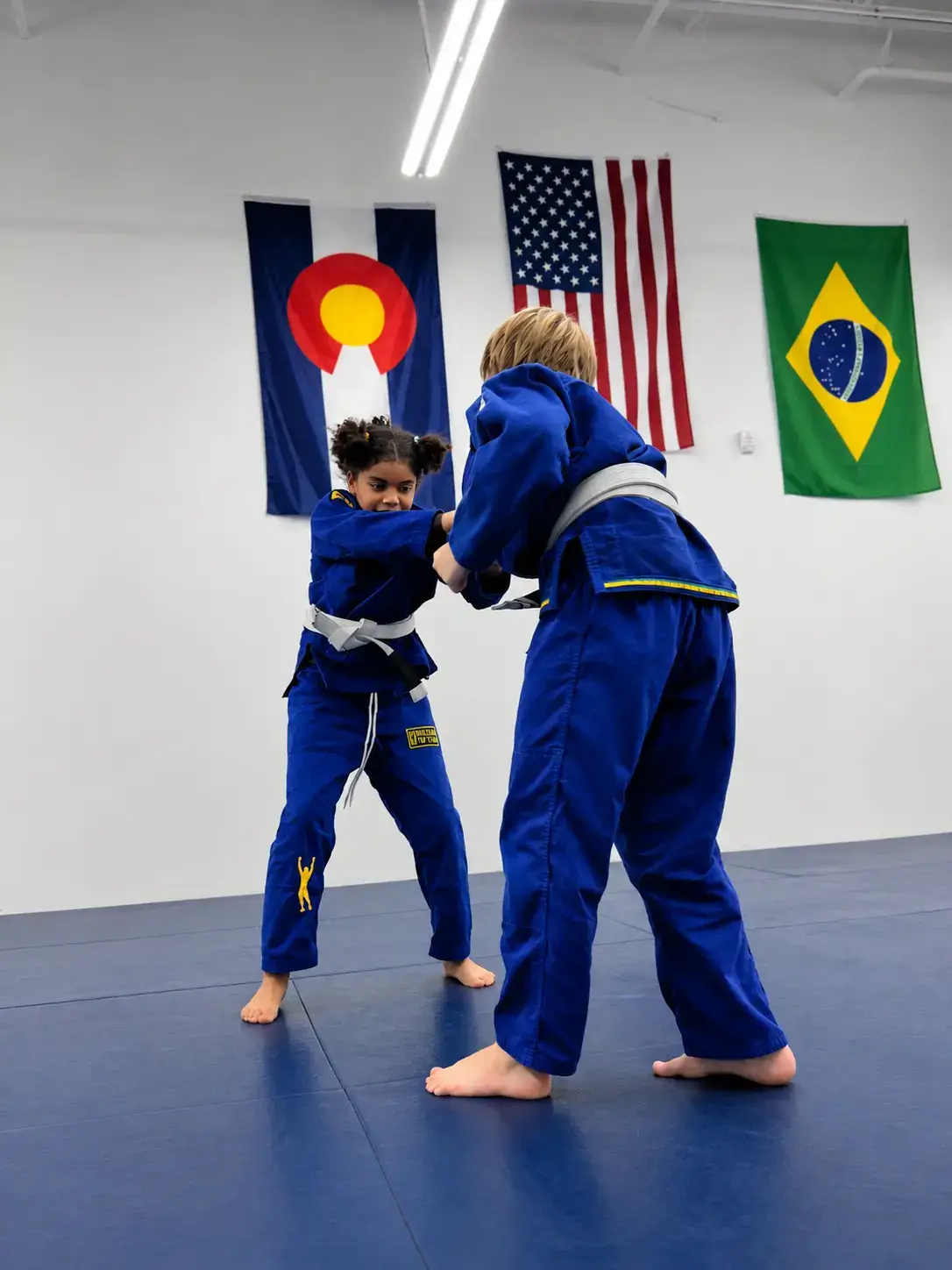 Two children in blue gis and white belts practice grappling on a blue mat.
