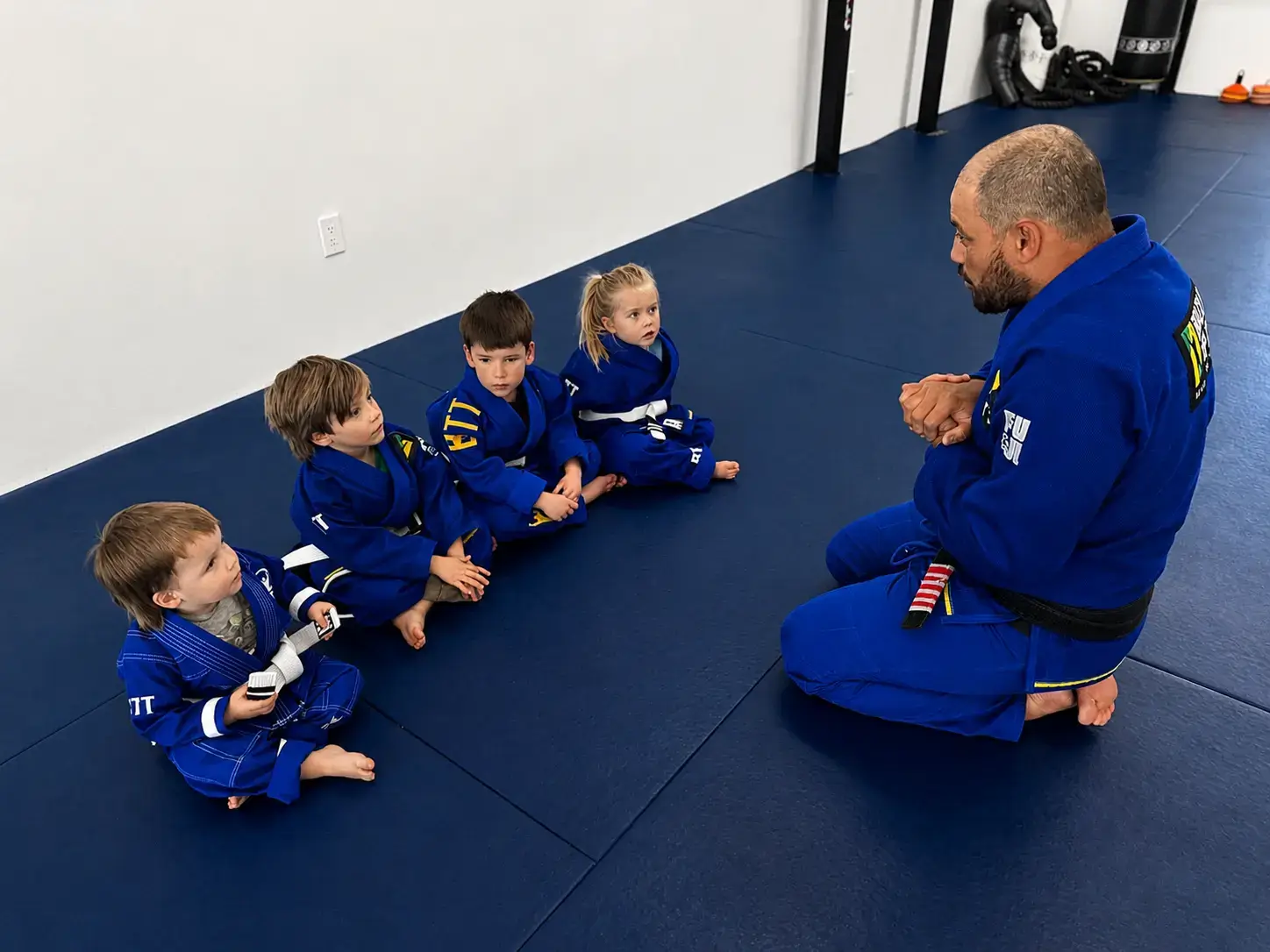 A black belt instructor kneels on a mat, teaching four young children in blue gis.