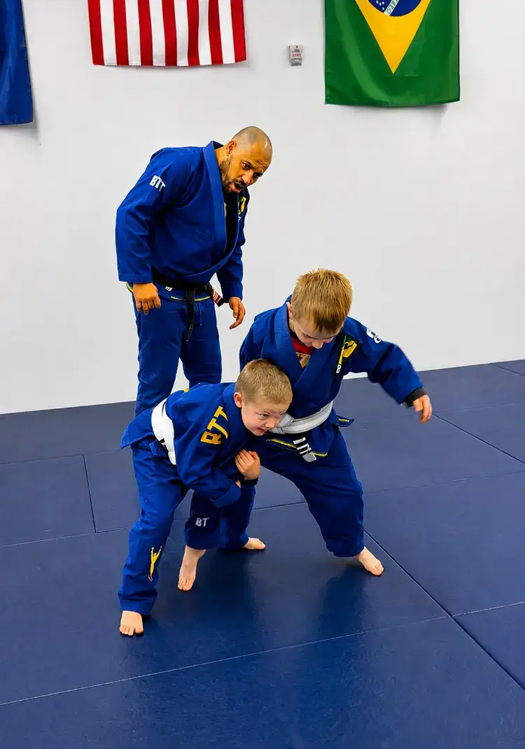 Two boys in blue gis practice Jiu-Jitsu techniques while a black belt instructor supervises.
