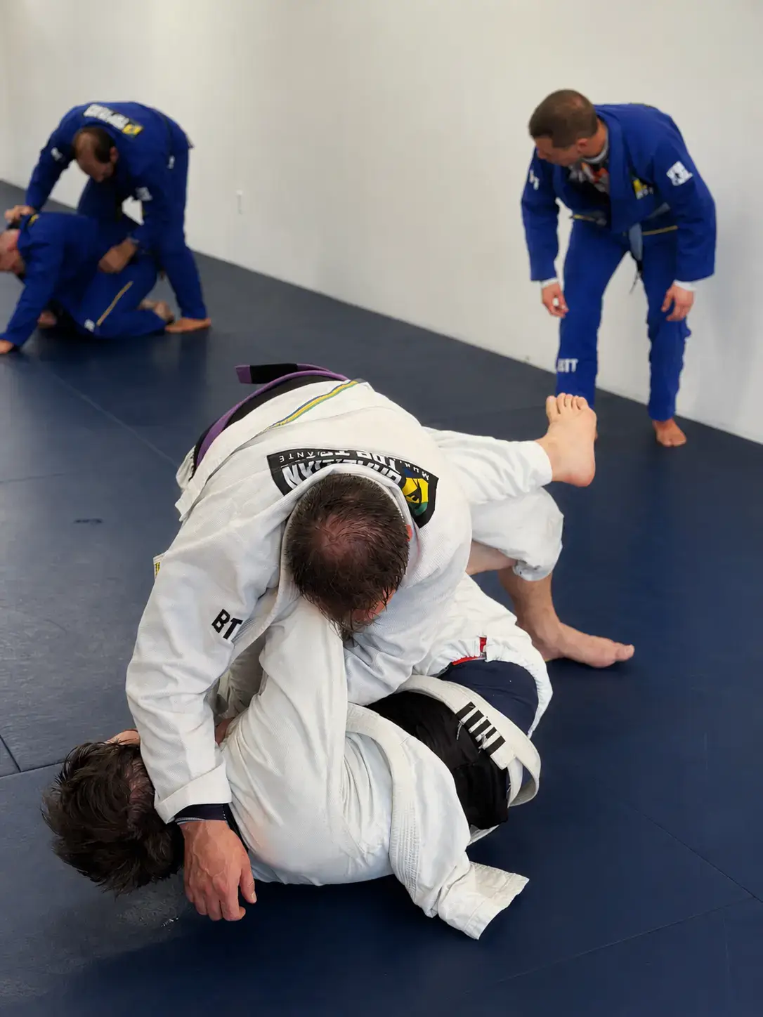 Men in white and blue gis practice Brazilian Jiu-Jitsu grappling on a blue mat.