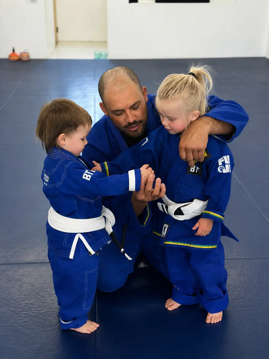 Male instructor in blue gi teaching two young children in blue jiu-jitsu uniforms on mats.
