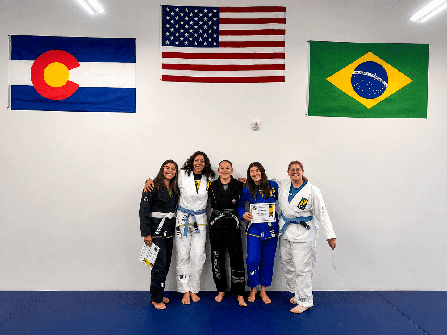 Five women in Jiu-Jitsu gis pose under Colorado, US, and Brazilian flags.