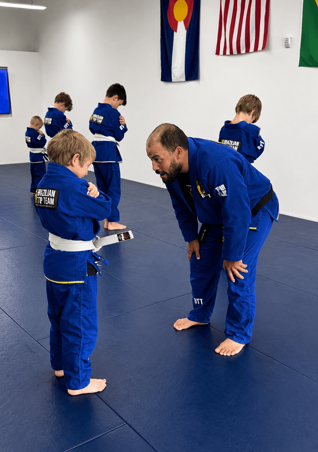 BJJ instructor leans down to talk to a young white belt student on blue mats.
