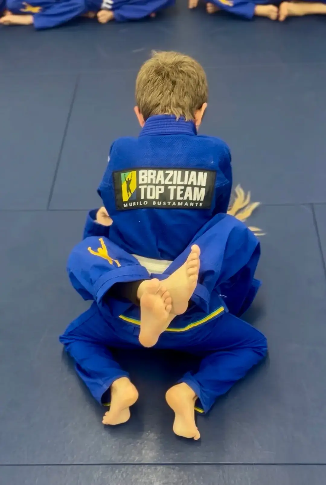 Two children wearing blue gis practice Brazilian Jiu-Jitsu grappling techniques on a blue mat.