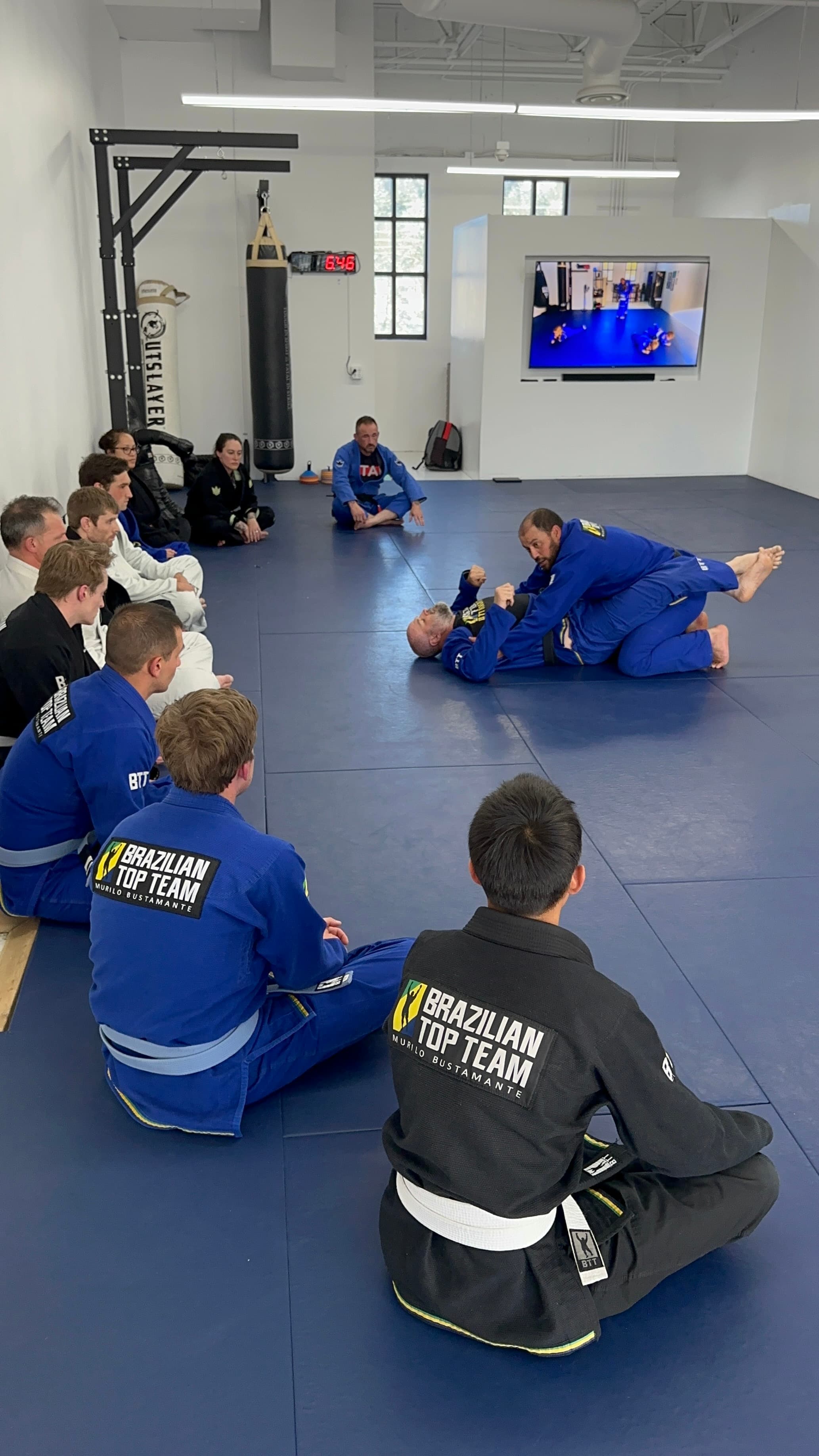 Brazilian Top Team students watch a Jiu-Jitsu demonstration on blue mats in a gym.