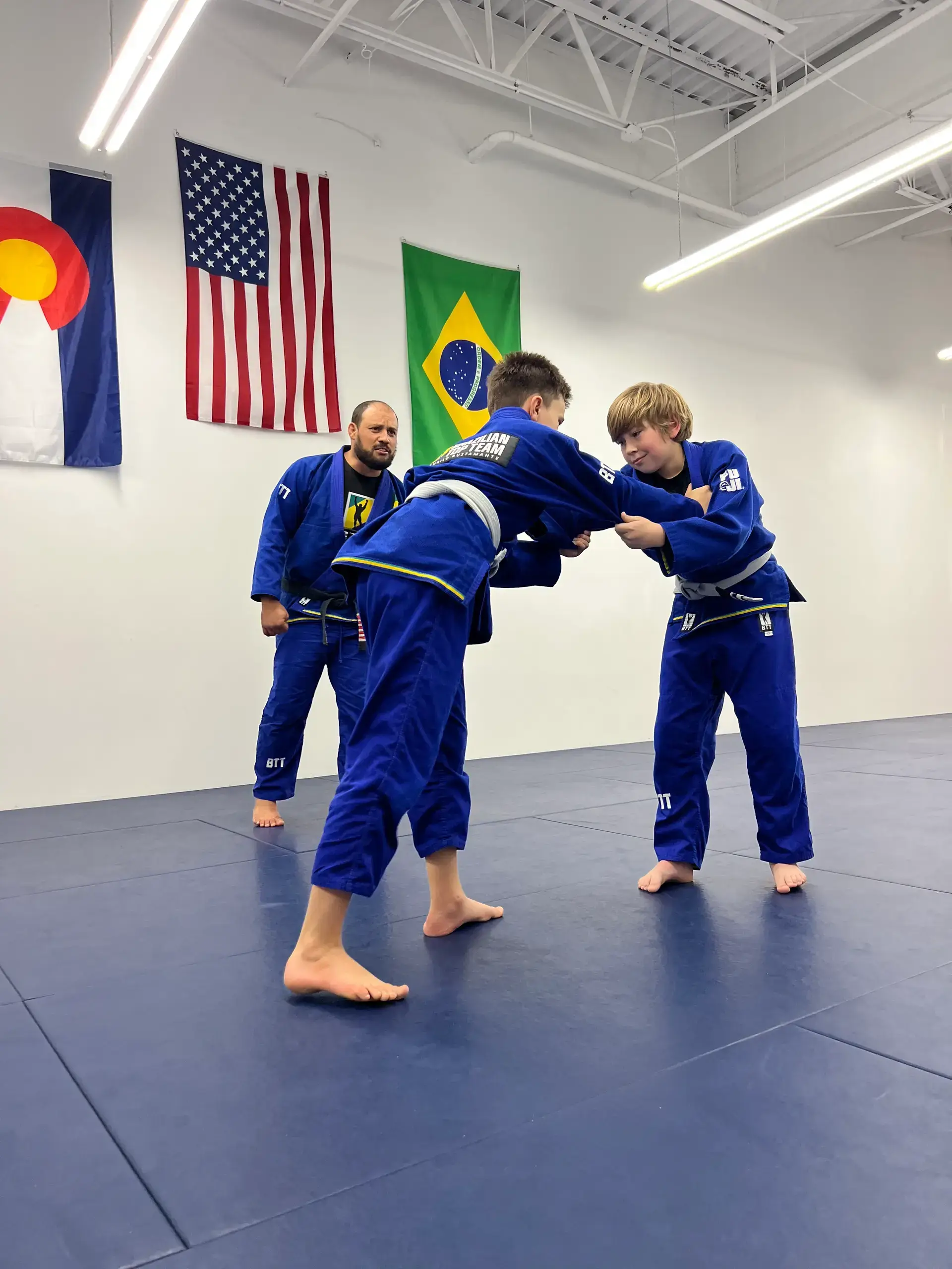 Two boys in blue gis practice Brazilian Jiu-Jitsu while an instructor watches them.