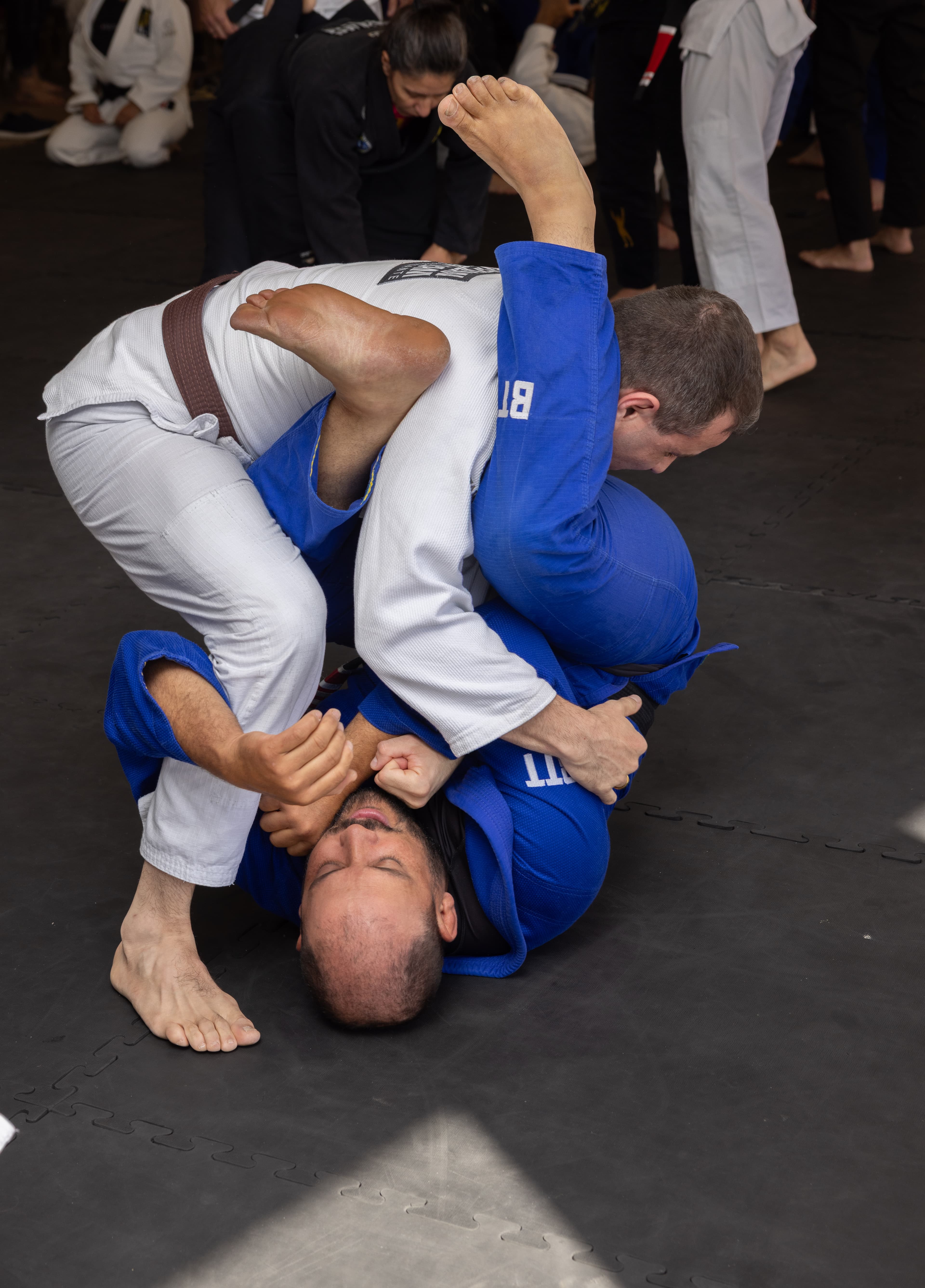 Brazilian Jiu-Jitsu practitioners in white and blue gis grappling on a black training mat.