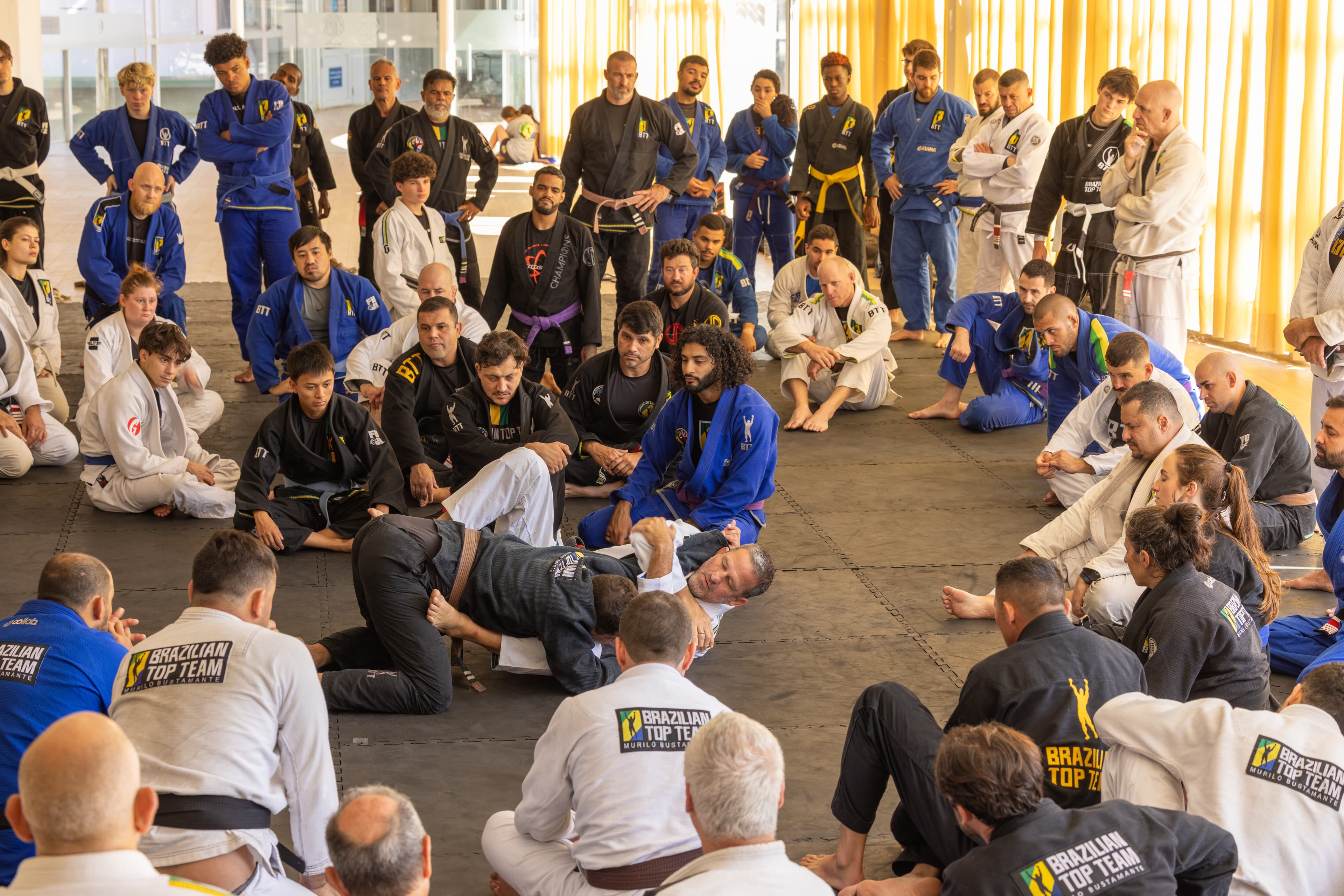 Brazilian Jiu-Jitsu practitioners in gis circle around two men demonstrating a grappling technique.